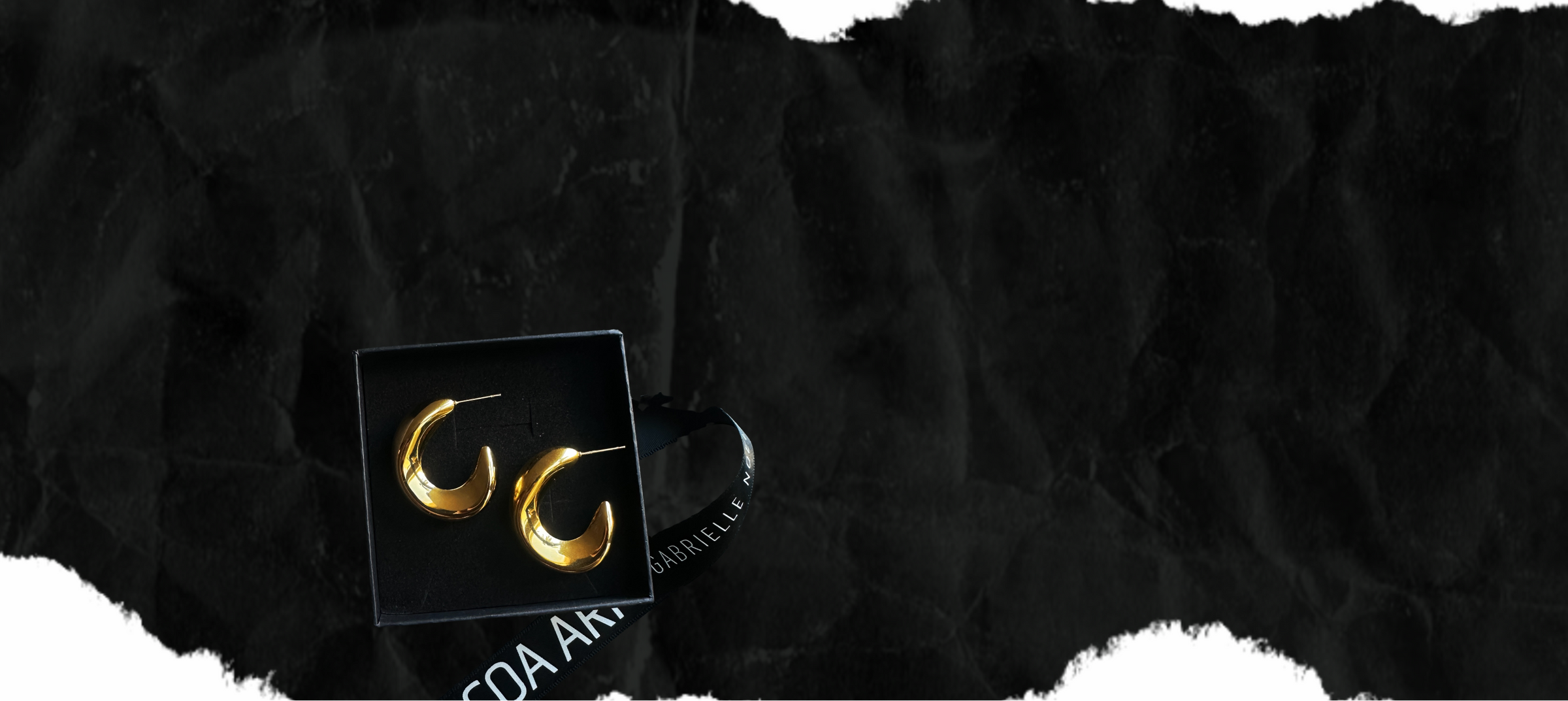 Gold earrings in a black box on a textured black background with cocoa ari black ribbon 