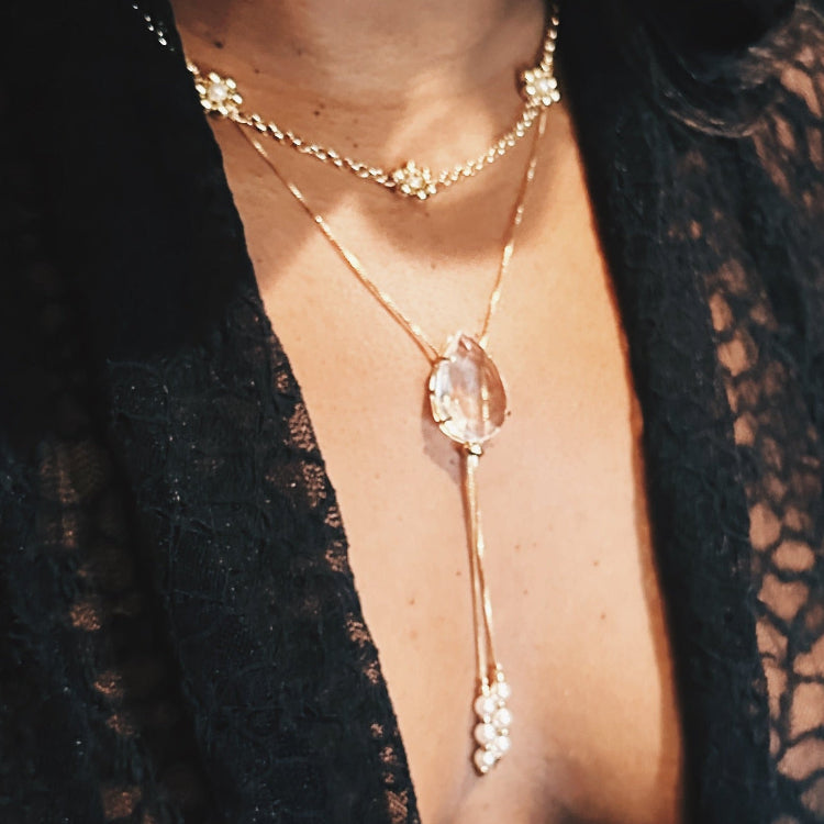 Close-up of a person wearing a delicate gold necklace with a clear stone pendant against a dark background.
